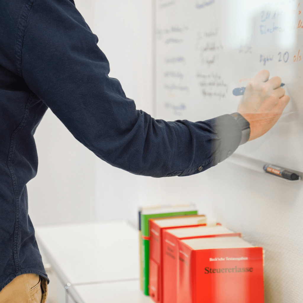 Arm und Hand einer Person, die etwas an ein Whiteboard schreibt Darunter mehrere Bücher, das vorderste trägt den Titel Steuererlasse.