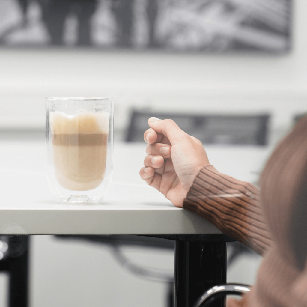 Ein Glas Latte Macchiato auf einem Tisch, daneben die Hand einer Mitarbeiterin. Verschwommener Hintergrund zeigt den Besprechungsraum.