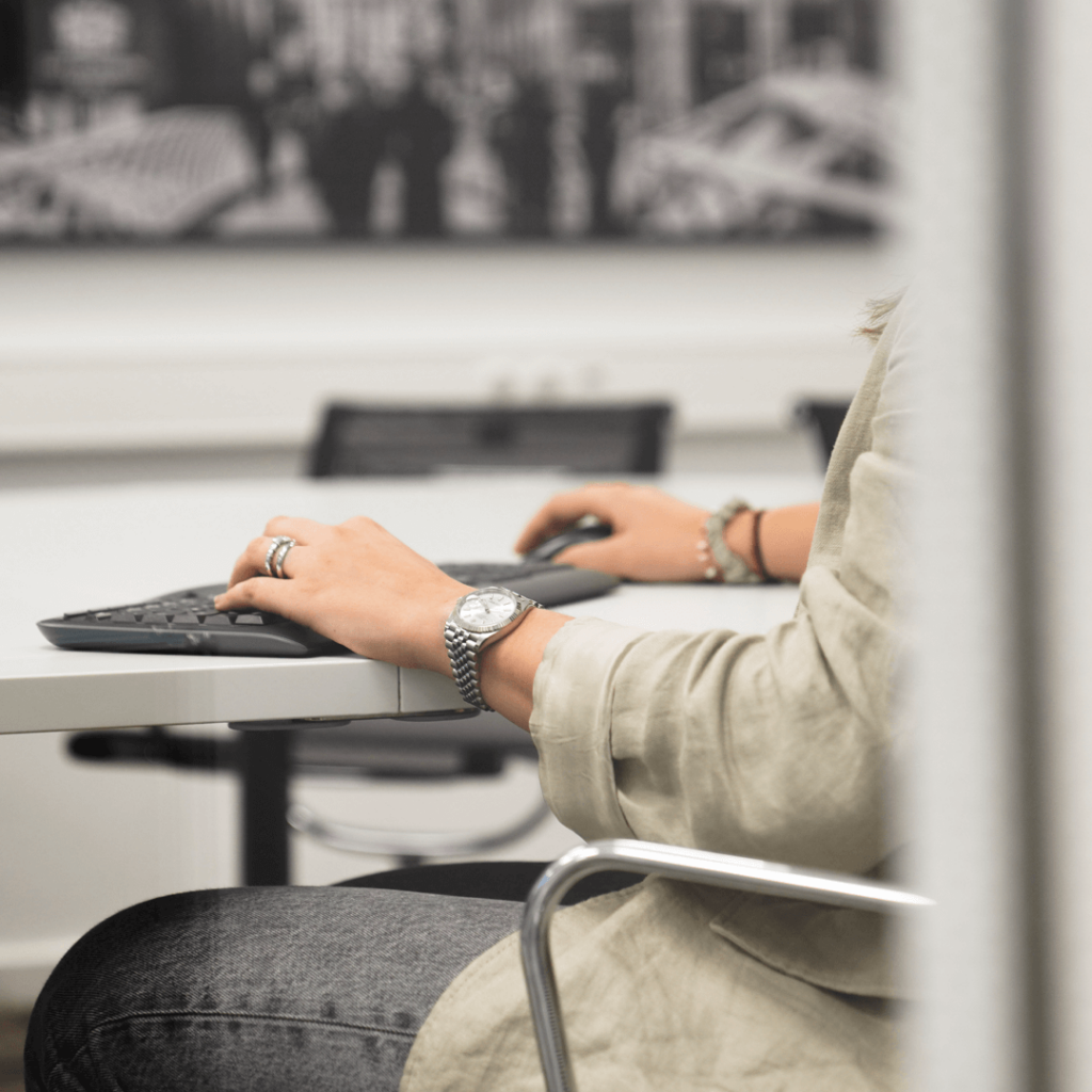 Person bei der Arbeit am Tisch, sichtbar sind nur ein Bein und zwei Hände an Tastatur und Maus