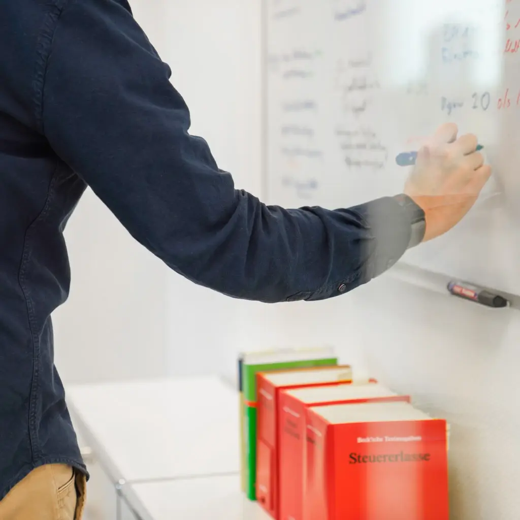 Arm und Hand einer Person, die etwas an ein Whiteboard schreibt Darunter mehrere Bücher, das vorderste trägt den Titel Steuererlasse.